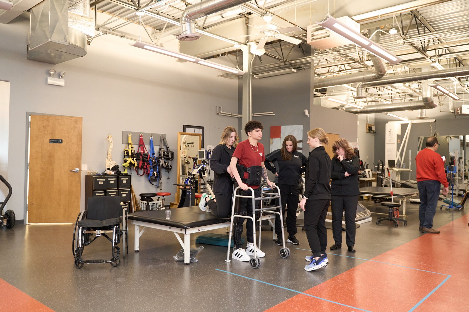 A young male patient with a back brace practices walking with a front-wheeled walker during a one-on-one rehabilitation session at Next Steps Chicago. A female therapist stands closely on his left to assist him, while three other staff members observe and monitor his progress. An empty wheelchair sits nearby on the left, and the background shows a spacious, well-lit neurological rehabilitation facility equipped with various physical therapy tools, exercise tables, and overhead tracking systems.